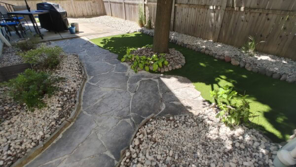 A backyard with a stone path, gravel landscaping, greenery around a tree, artificial grass, and a wooden fence. Grill and table are visible on a stone patio in the background.