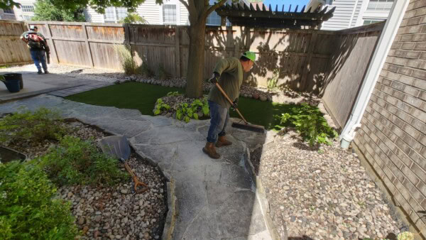 A person in work clothes uses a broom to sweep a stone pathway in a backyard garden, while another person stands in the background near a wooden fence and artificial grass.