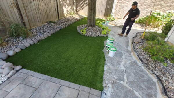 A person stands on a stone path next to a yard with artificial grass, paint buckets, a tree surrounded by rocks, and garden beds with stones along a wooden fence.