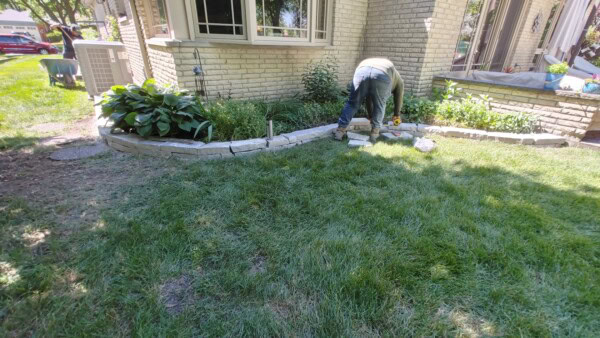 A person is working in a garden by a house, placing stone bricks to create a curved border around a flower bed. Green grass covers the yard and leafy plants are along the edge.