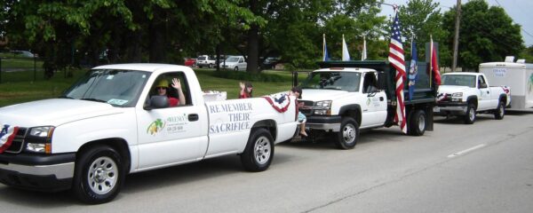 A parade with decorated vehicles, including a white pickup truck with a Remember Their Sacrifice sign, American flags, and people waving, moves down a street lined with trees.