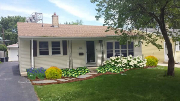 A small beige house with a tan roof, surrounded by a neat front yard featuring green plants and flowering bushes, labeled with plant names, and a stone path leading to the front door.