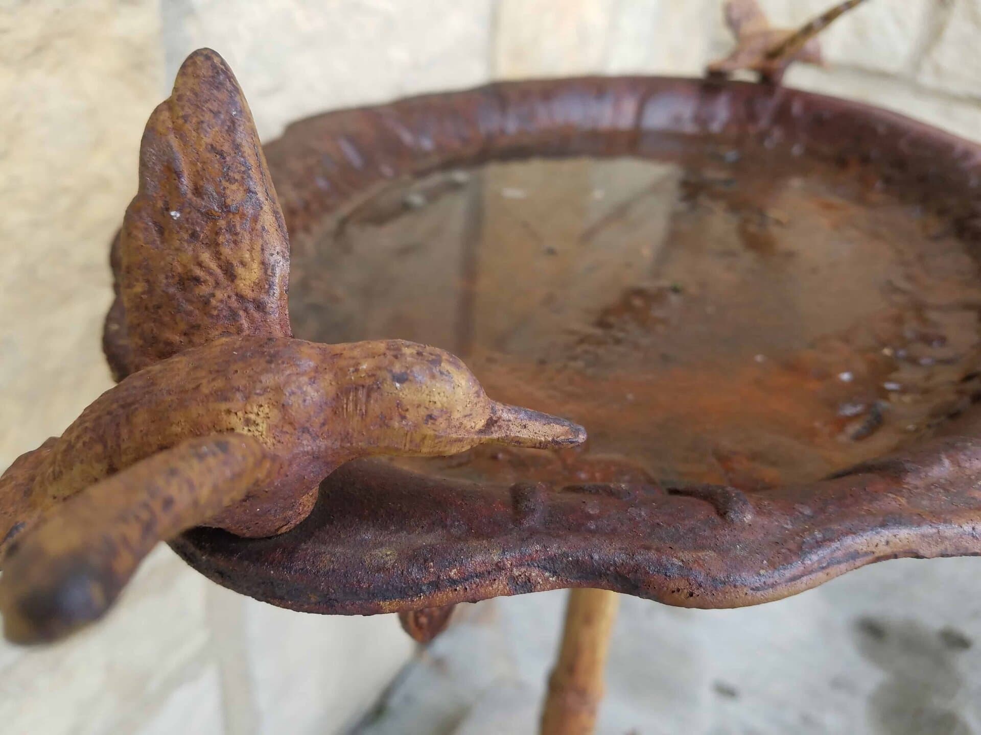 A close-up of a rusted metal bird sculpture attached to the rim of a weathered birdbath filled with water. The background is a blurred stone surface.