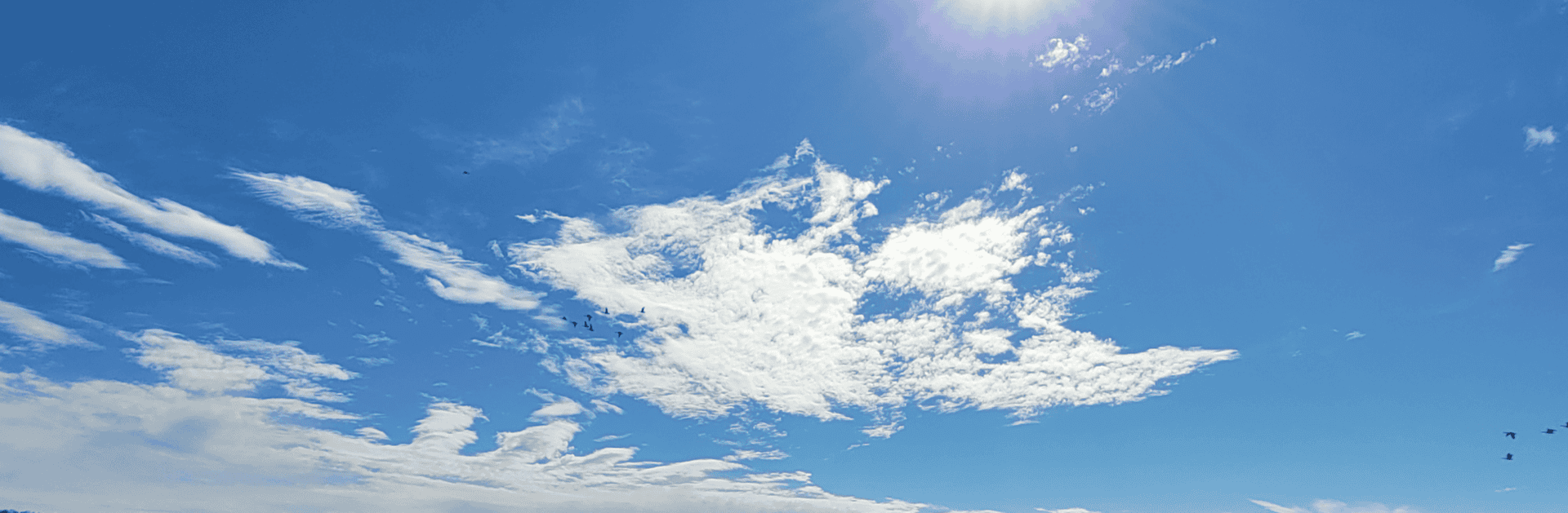 Bright blue sky with scattered white clouds and the sun shining at the top right. A few birds are visible flying in the distance near the upper right edge.