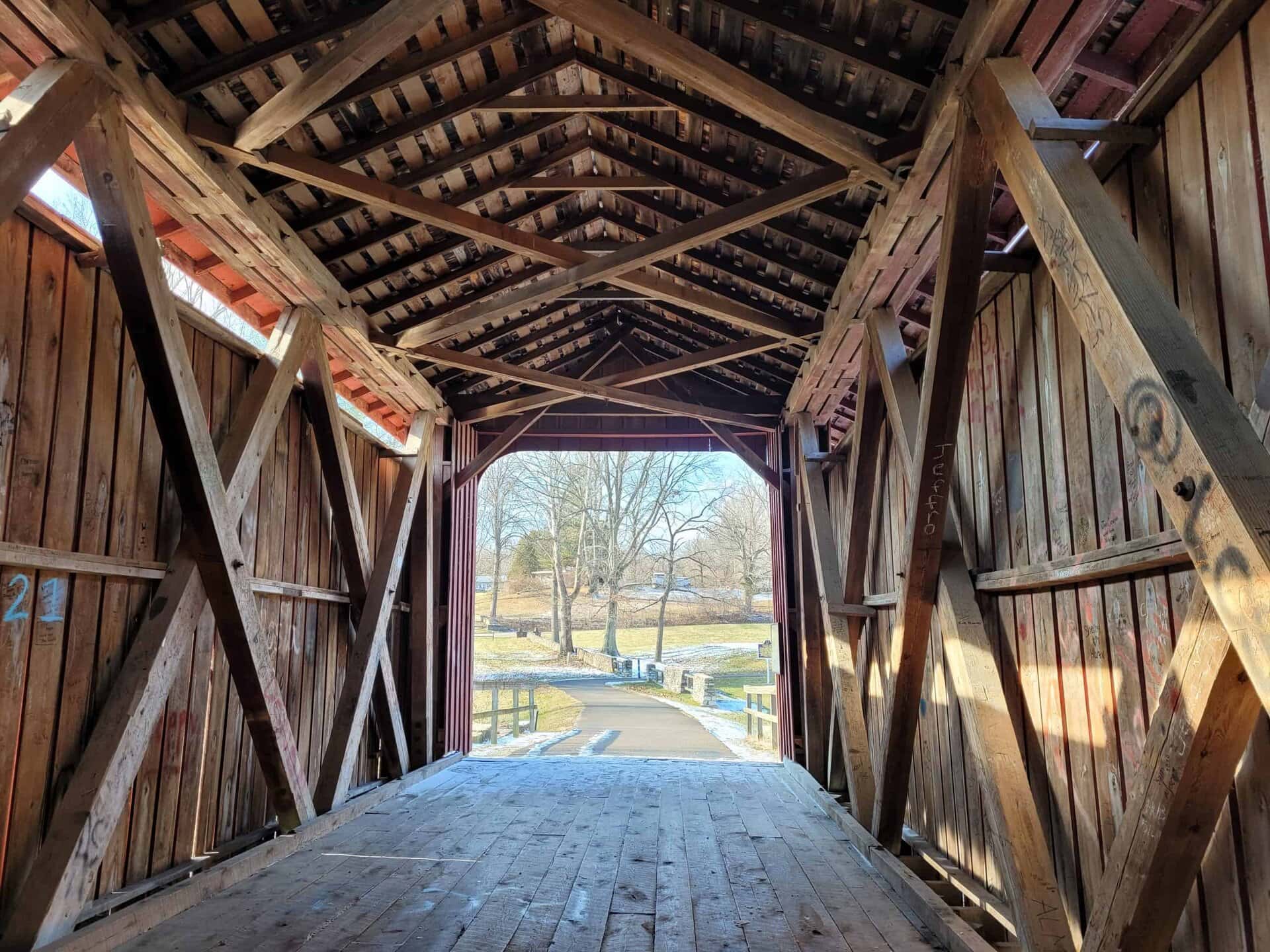 View from inside a wooden covered bridge, looking out toward a sunny landscape with trees, grass, and a paved path in the distance.