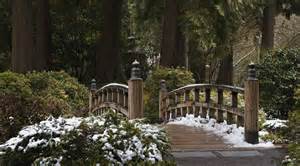 A small wooden bridge crosses over a path in a wooded area, with patches of snow covering the ground and surrounding green bushes.