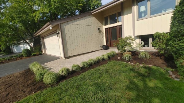 A modern beige house with a double garage, landscaped front yard, paved walkway, ornamental grasses, small shrubs, and green lawn under leafy trees.