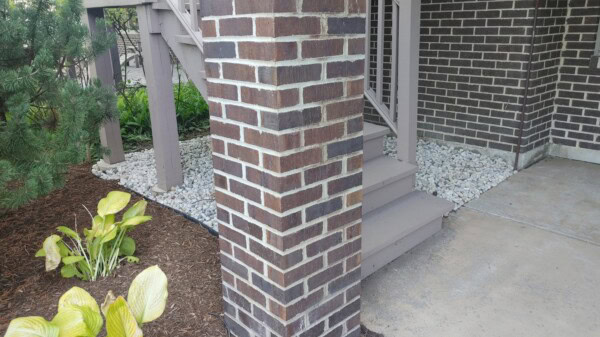 A brick pillar stands near stairs leading to a porch. White gravel borders the building, and mulch with green and yellow plants is in the foreground. A concrete walkway is on the right.