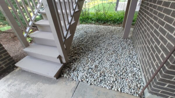 Stairs with light-colored wooden steps lead to an upper level. Beneath the stairs, a section of ground is covered with small white pebbles next to a brick wall and some green plants.