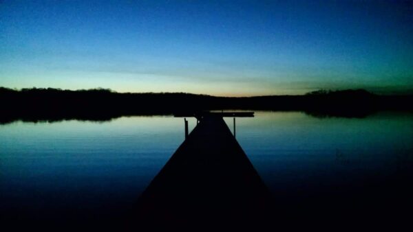A wooden dock extends into a calm lake at dusk, with a dark treeline in the background and a gradient sky shifting from deep blue to pale yellow near the horizon.