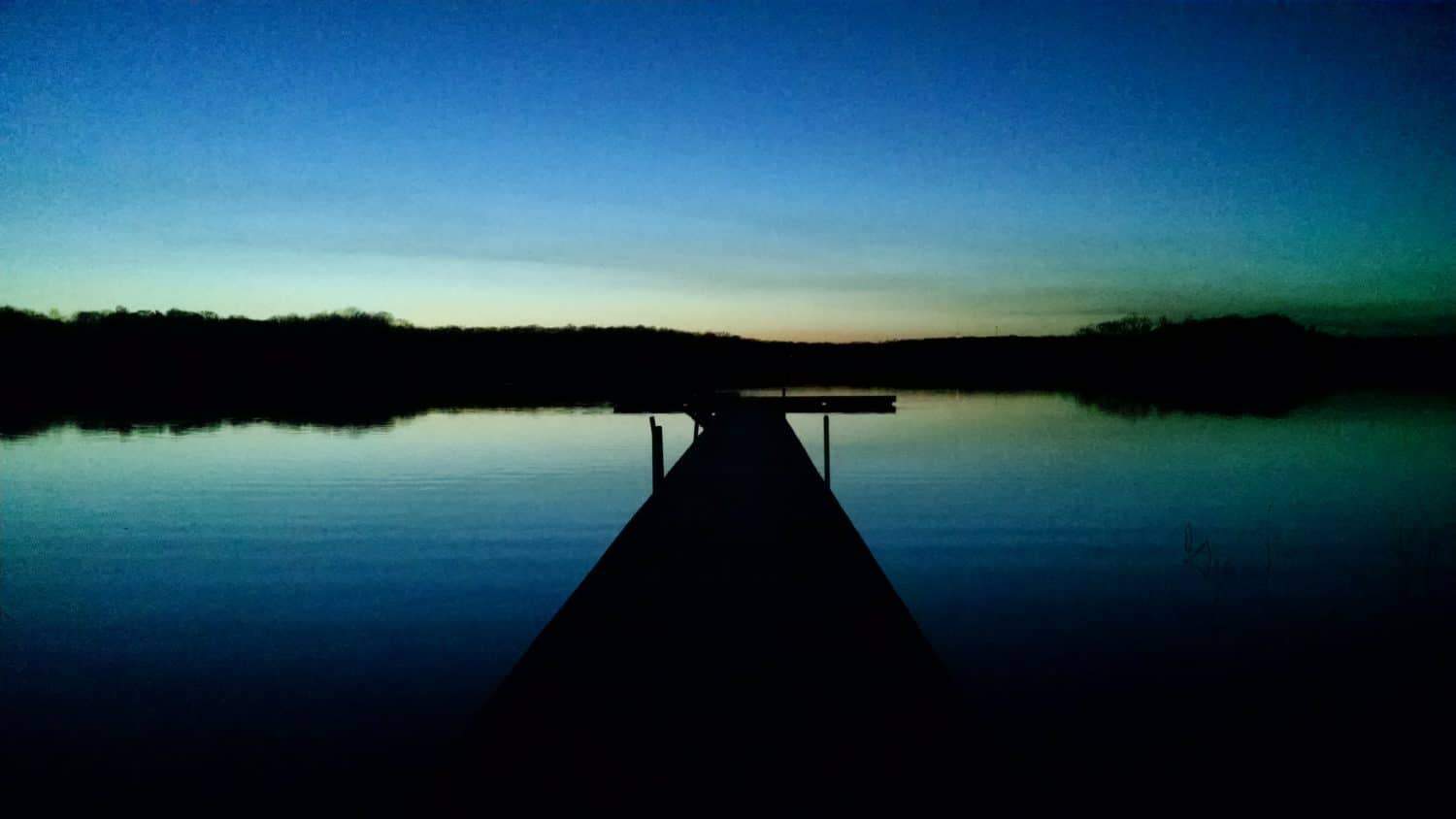 A wooden dock extends into a calm lake at dusk, with a dark treeline in the background and a gradient sky shifting from deep blue to pale yellow near the horizon.