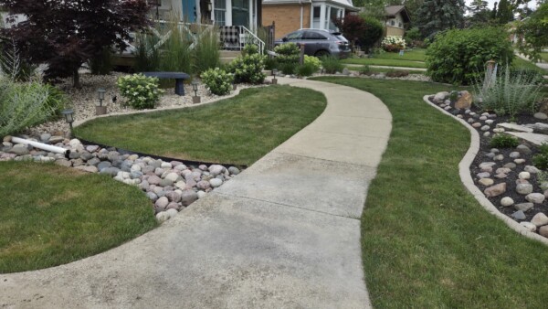 A curved concrete walkway winds through a neatly landscaped front yard with green grass, flower bushes, small rocks, shrubs, and a parked car in the driveway.
