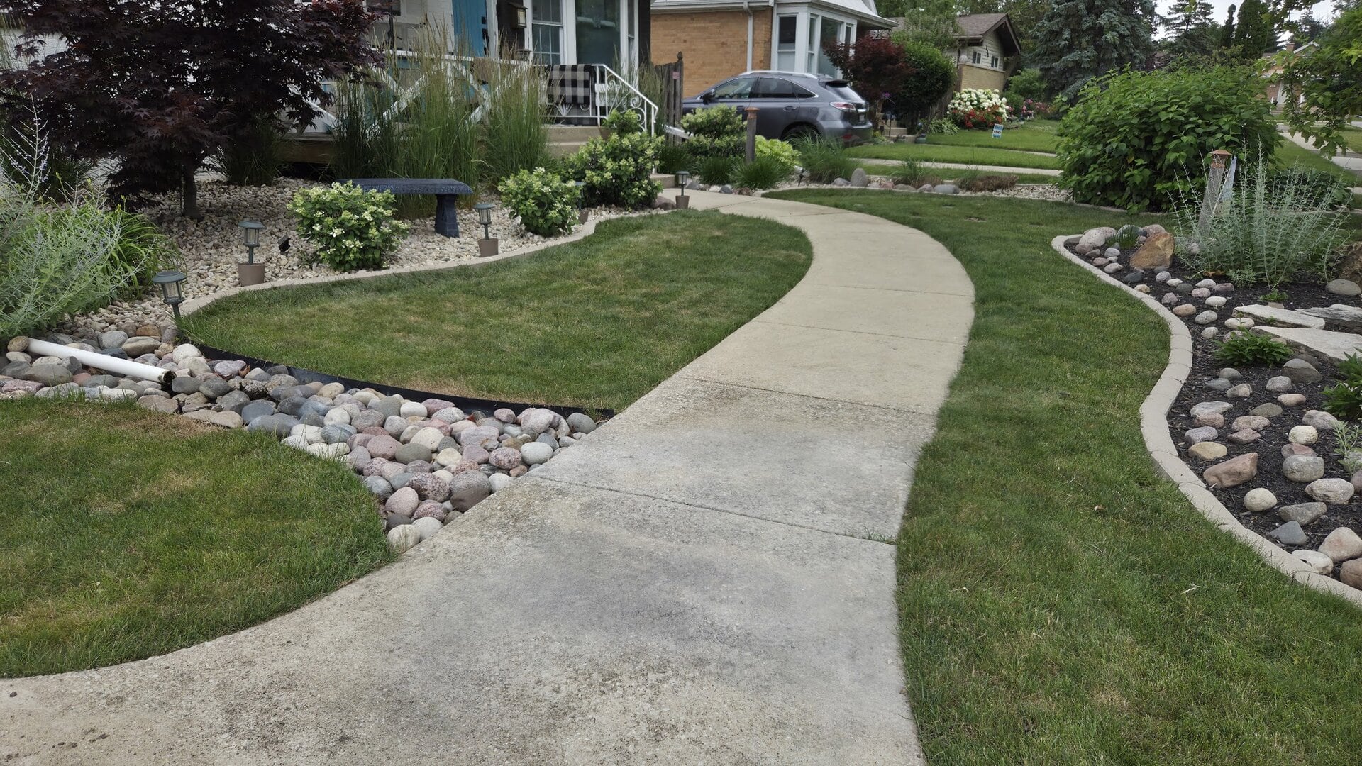 A curved concrete walkway winds through a neatly landscaped front yard with green grass, flower bushes, small rocks, shrubs, and a parked car in the driveway.
