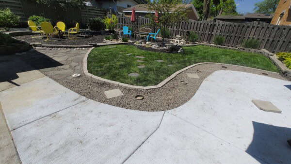 A backyard with a curved concrete walkway, gravel, stone path, and two seating areas—one with yellow chairs, one with blue—surrounded by grass, plants, and a wooden fence under a clear sky.