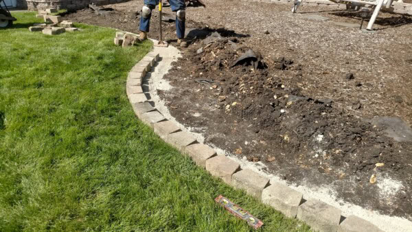 A person builds a curved stone border between a grassy lawn and a dirt garden bed. Bricks are stacked in a line, and a level tool lies on the grass nearby.