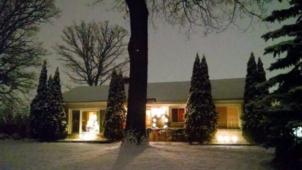A house decorated with holiday lights and a wreath on the front door, surrounded by tall evergreen trees and snow covering the ground at night.