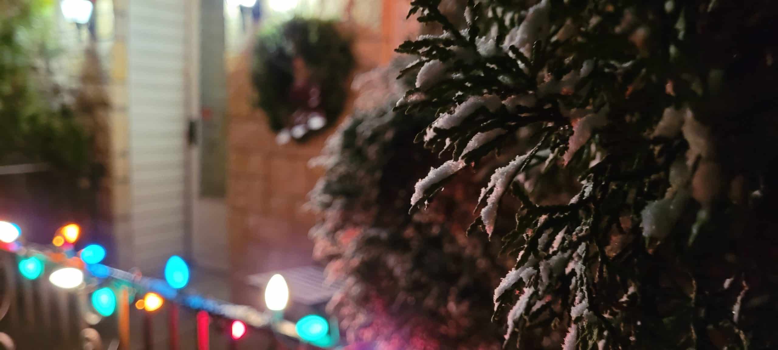 Snow-covered evergreen branches in the foreground with colorful holiday lights and a wreath on a door in the blurry background, creating a festive winter scene at night.