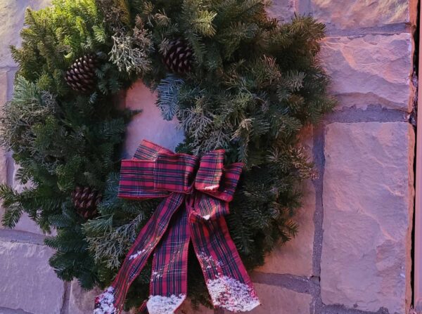 A green wreath decorated with pinecones and a red plaid bow, hanging on a stone wall. Some of the bows ribbon ends have artificial snow.