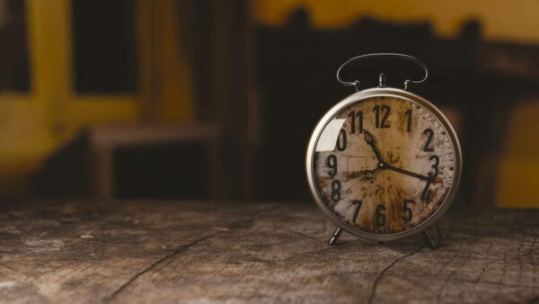 A vintage alarm clock with a worn face sits on a rustic wooden surface, showing the time as 10:11. The background is blurred and dimly lit.