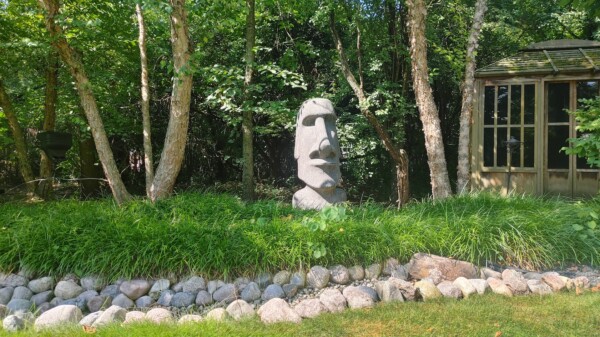 A stone Easter Island-style head statue stands among tall grass and trees, with a rock border and a wooden gazebo nearby on a sunny day.