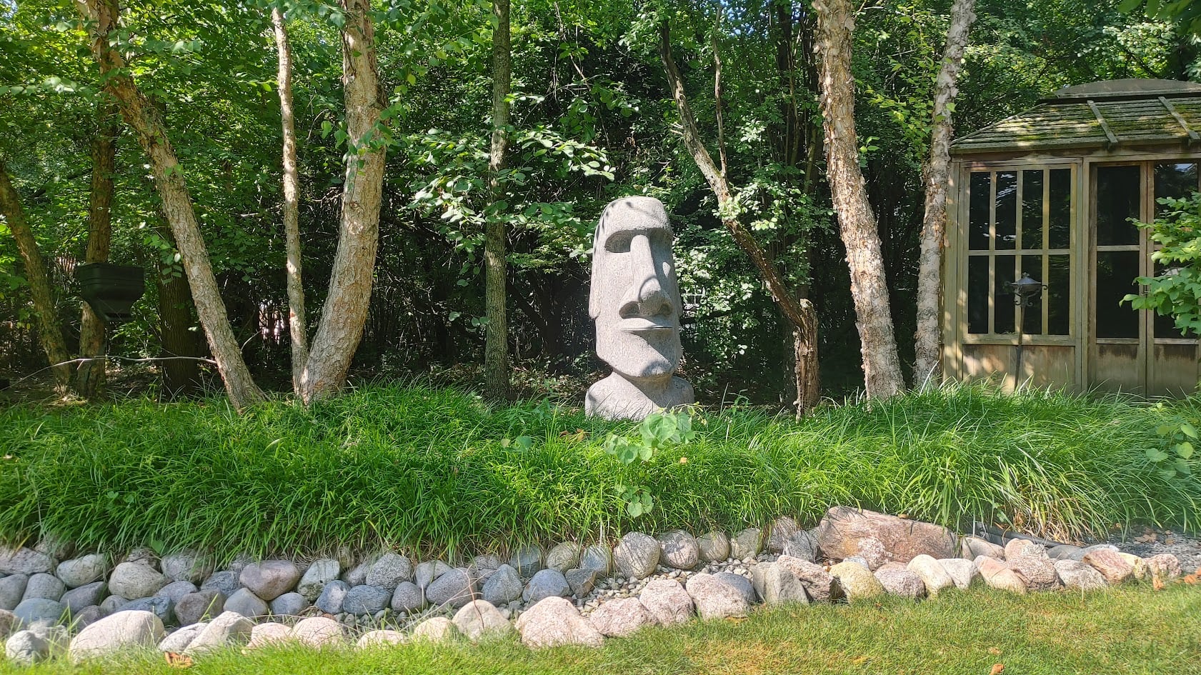 A stone Easter Island-style head statue stands among tall grass and trees, with a rock border and a wooden gazebo nearby on a sunny day.