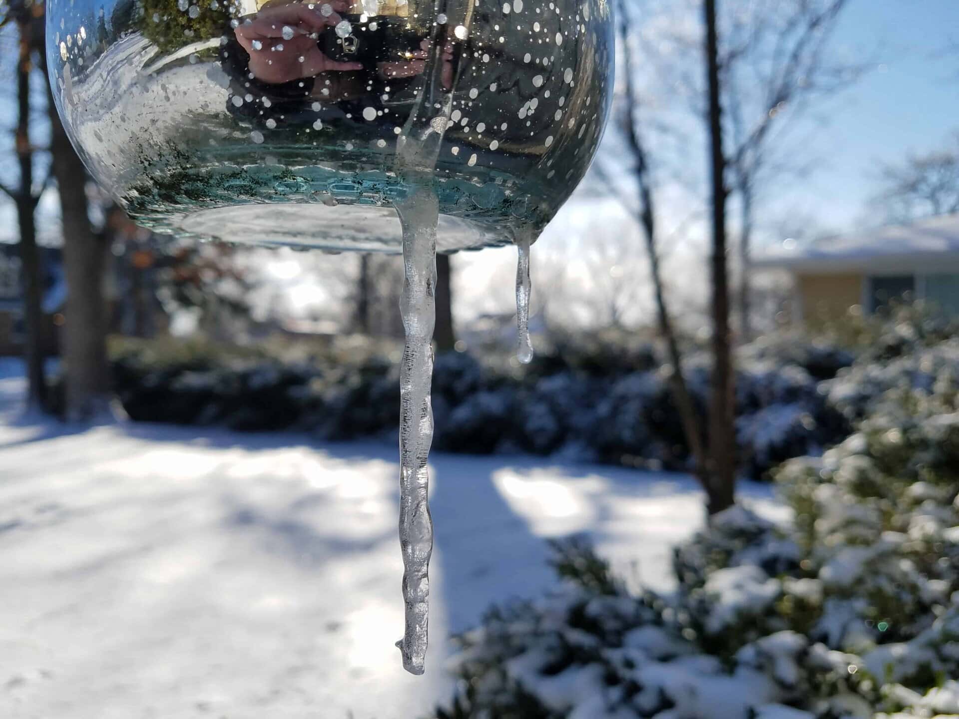 Close-up of a glass object with icicles hanging from it. Snow covers the ground and trees in the background, with a house partially visible on the right. Sunlight creates reflections on the glass.