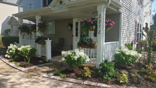 A house with a covered porch, hanging flower baskets, white blooming bushes, and a stone-bordered garden. The house number 2372 is on a porch post. Sunlight casts shadows on the sidewalk and yard.