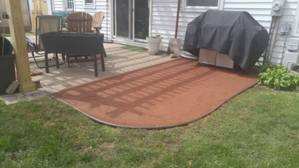 A backyard patio with chairs, a table, a covered grill, and a curved patch of newly spread reddish-brown dirt bordered by black edging next to green grass.