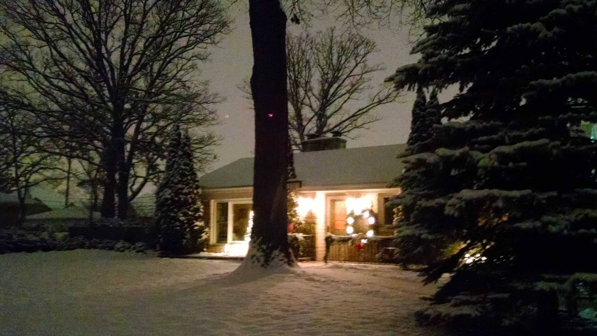 A cozy house glows with warm lights on a snowy night, surrounded by snow-covered trees and a fresh blanket of snow on the ground.
