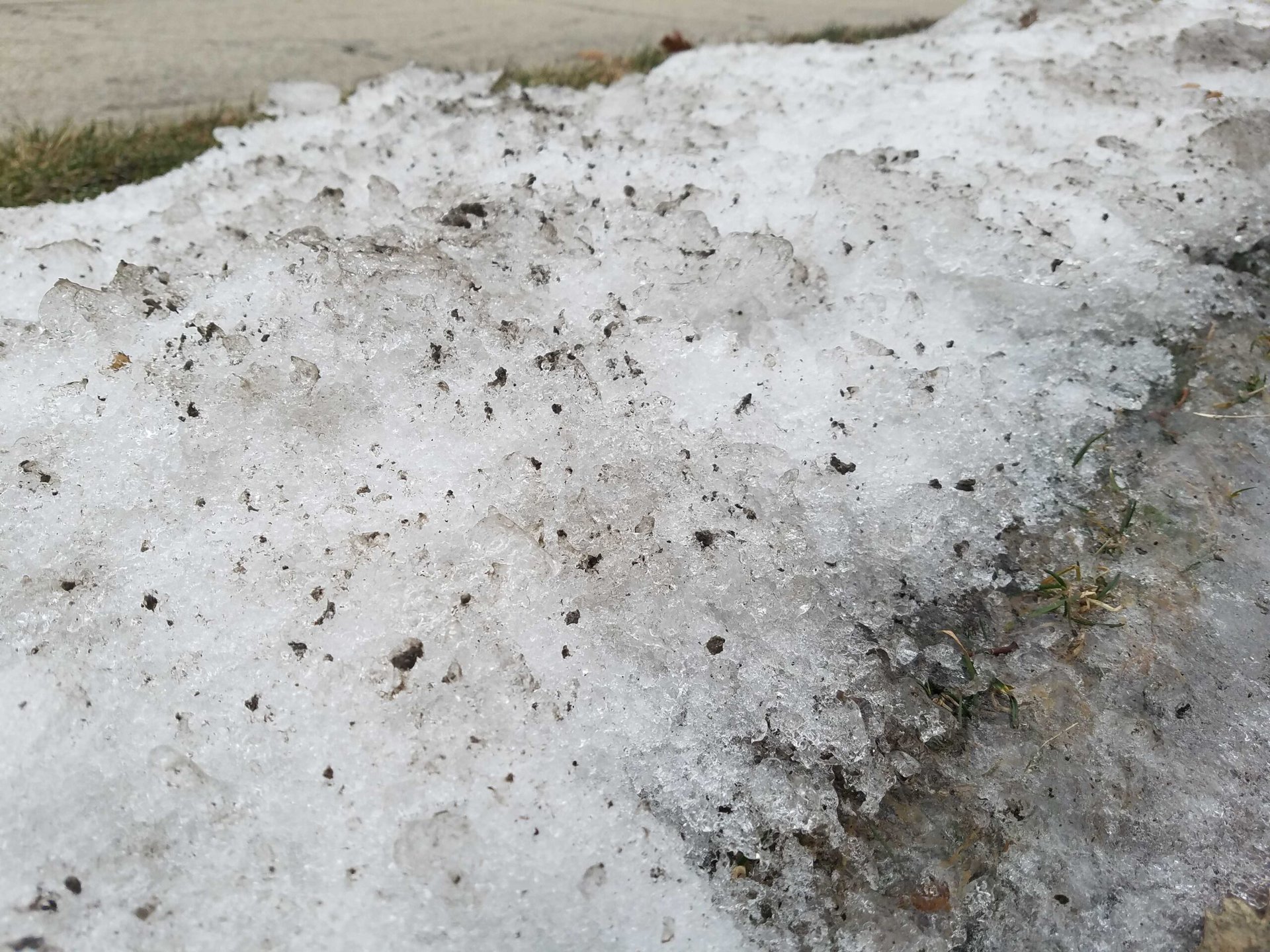 Close-up of dirty, melting snow on the ground with some grass peeking through, next to a road.