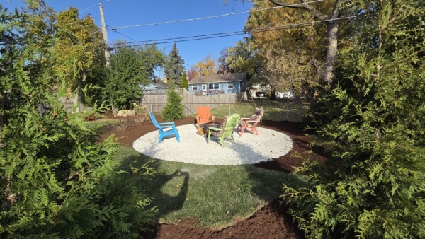 Four colorful Adirondack chairs surround a fire pit on a gravel circle in a backyard. Tall green shrubs line the edge, and a house with blue siding is visible in the background. Bright sunlight casts shadows.
