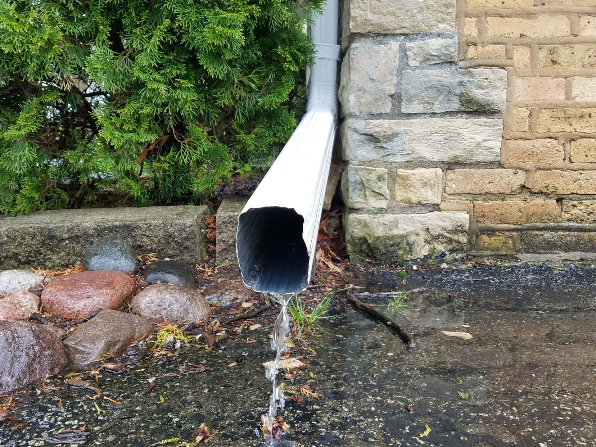 A close-up of a white downspout with water flowing out, spraying onto wet pavement. Stones, a green shrub, and a brick wall are visible in the background.