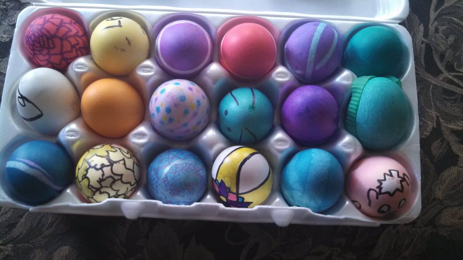 A carton with 18 colorful Easter eggs, each uniquely decorated with patterns, doodles, and vibrant solid colors, resting in a white egg tray on a dark patterned surface.