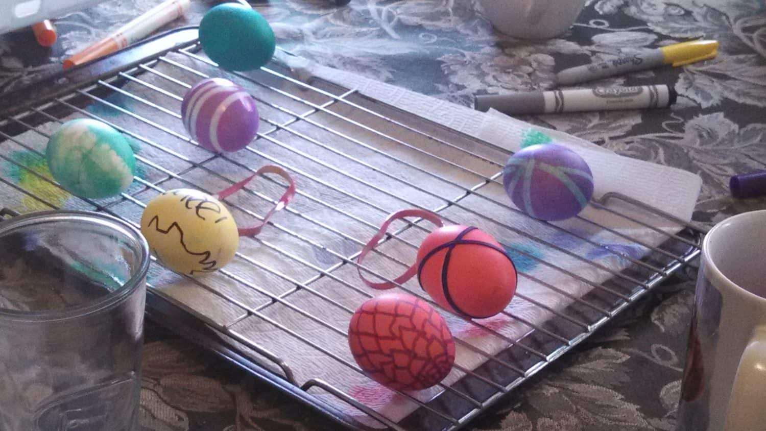 Several brightly colored and decorated Easter eggs rest on a cooling rack over a paper towel, surrounded by markers, a glass, and a mug on a patterned tablecloth.