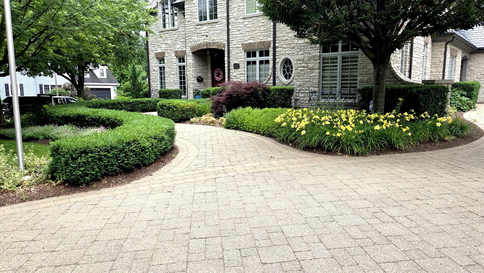 A stone house with large windows is behind a curved, paved driveway, surrounded by manicured hedges, green shrubs, and yellow flowers under leafy trees.