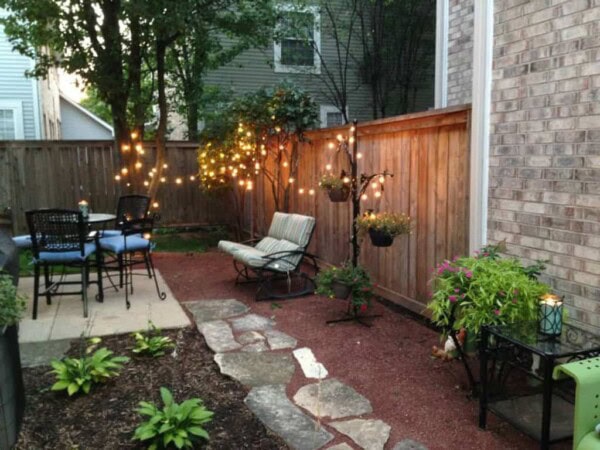 A cozy backyard patio with string lights, a cushioned chair, small table, potted plants, and a dining table with chairs. The space is bordered by a wooden fence and landscaped garden beds.
