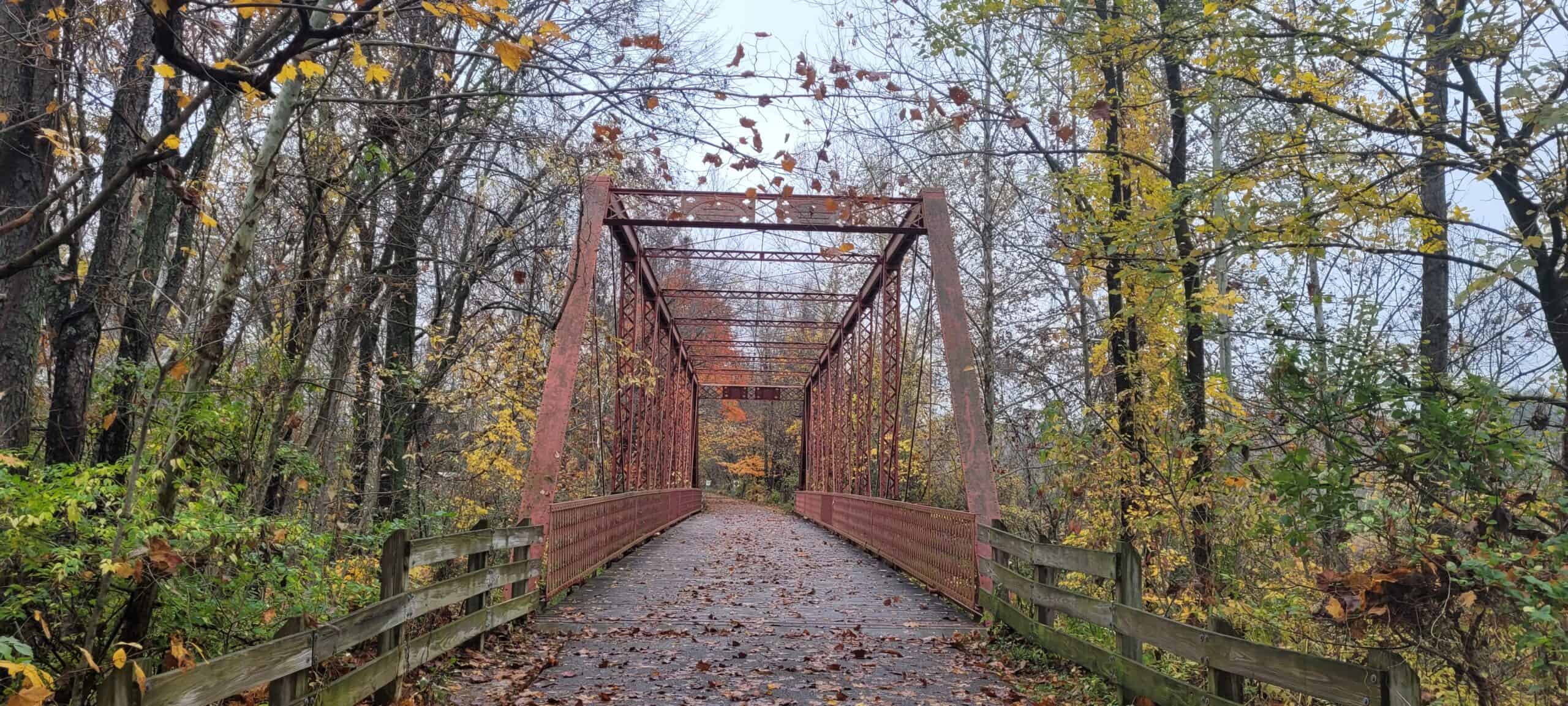 A rusty metal bridge with wooden railings crosses through a forest of autumn trees, leaves scattered on the path and branches overhead.