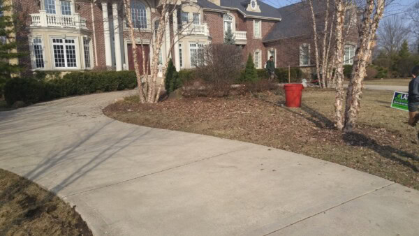 A curved driveway leads to a large brick house with tall windows. Bare trees and bushes line the yard. A red bin sits on the lawn, and a person stands near a green sign by the sidewalk.