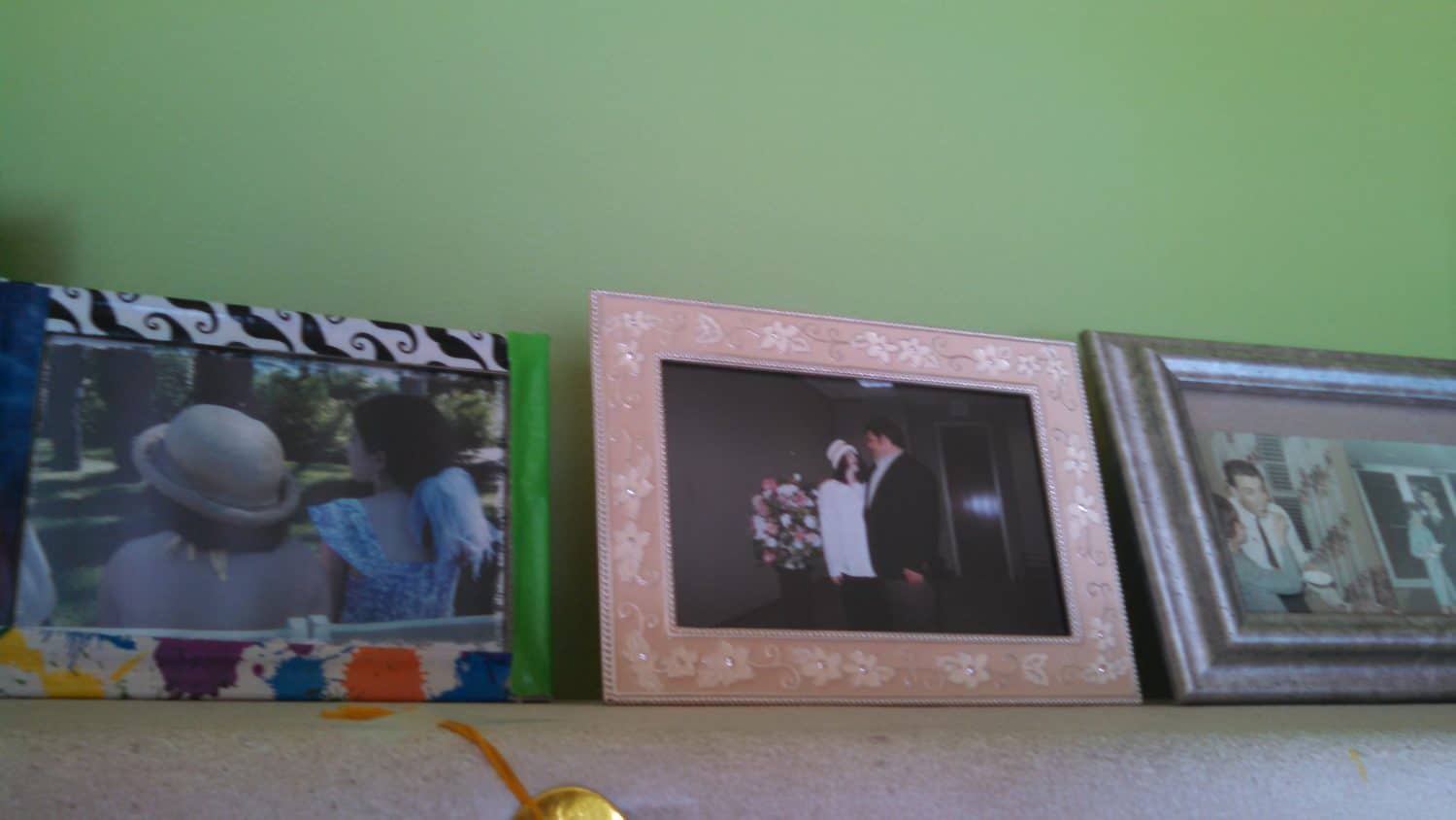 Three framed photos on a shelf: two people outdoors, a couple at a wedding, and a solo person. All frames are different styles; the background wall is green.