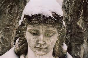 A weathered stone statue of an angel with a serene expression is dusted with snow, highlighting its hair and face.