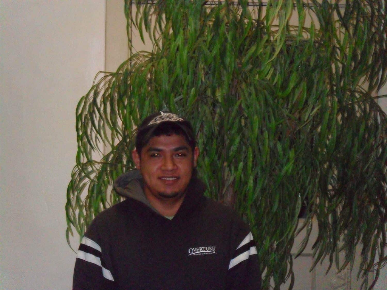 Fernando B. A person wearing a dark hoodie and a cap stands smiling in front of a large leafy indoor plant against a cream-colored wall.