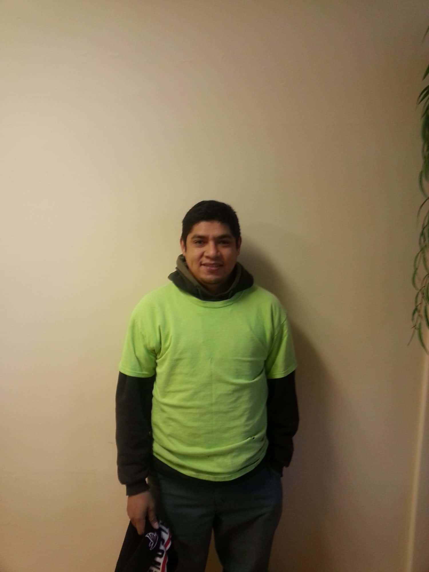 A person wearing a neon green T-shirt over a dark hoodie stands in front of a light-colored wall, holding a black item. Some green leaves are visible at the top right corner.