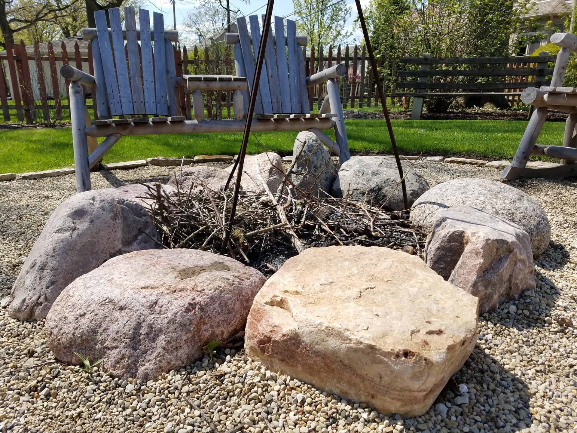 Large stones form a fire pit filled with twigs. Two wooden chairs and a bench sit in the background on a gravel surface, with green grass and a wooden fence behind them.