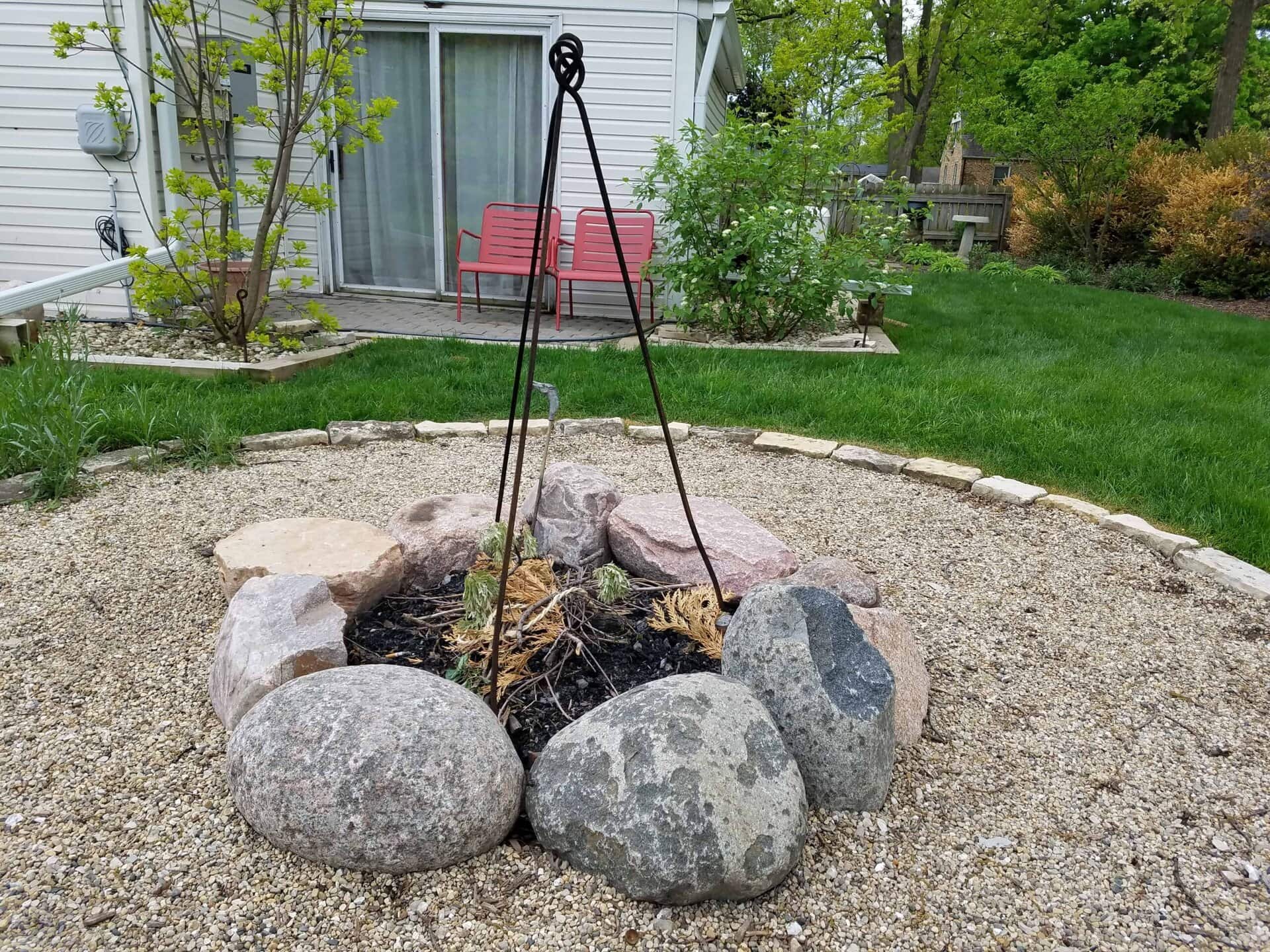 A circular fire pit made of large rocks sits on gravel, with a metal tripod over it. Behind, theres green grass, bushes, and a white house with red chairs on a patio near sliding glass doors.