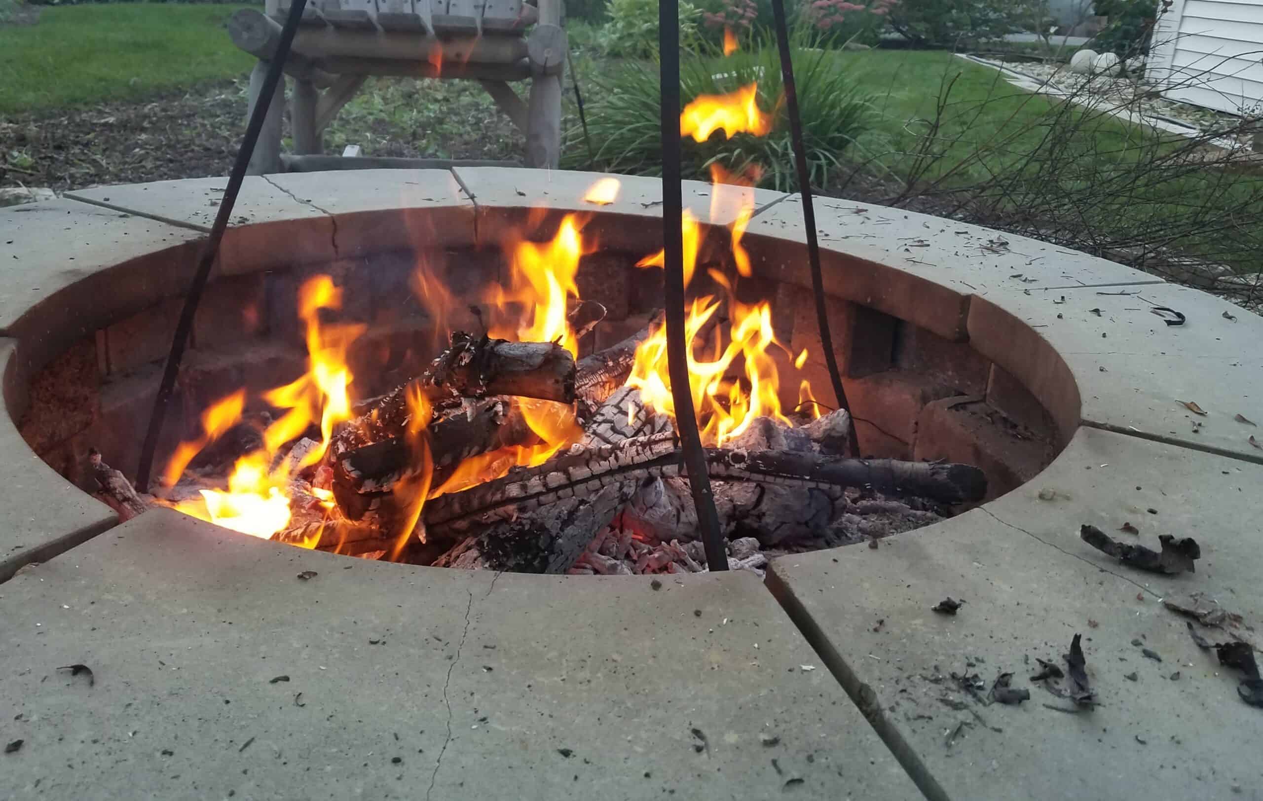 A close-up of a fire pit with burning logs and bright flames, surrounded by a stone ring. Grass, plants, and a house are visible in the background.
