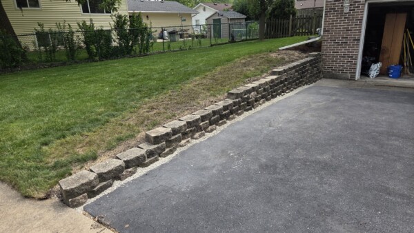 A paved driveway runs beside a short retaining wall of stacked concrete blocks, separating it from a grassy yard. The wall leads to a brick garage building in a suburban residential area.