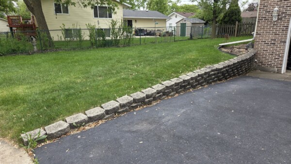 A small retaining wall made of stone blocks borders an asphalt driveway, separating it from a sloped grassy yard with houses and sheds in the background.