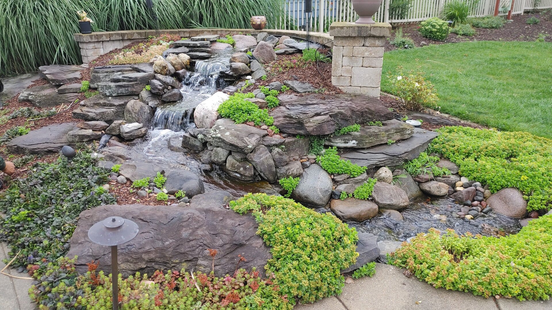 A small backyard waterfall flows over stacked rocks, surrounded by green ground cover and plants, with a grassy lawn and stone pathway nearby.