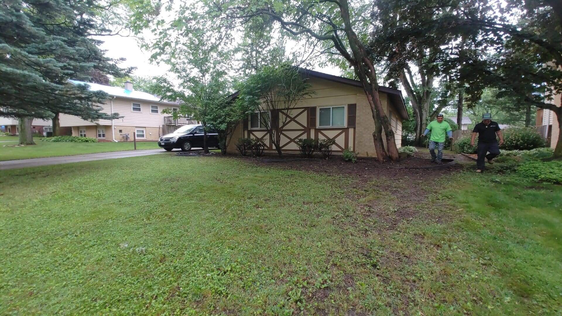 A person in a green shirt uses a string trimmer along the edge of a grassy yard next to a tan house with decorative wooden trim; a black vehicle is parked nearby.