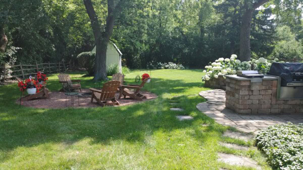 Sunny backyard with Adirondack chairs around a fire pit, surrounded by trees and flowers. A brick patio with a barbecue grill is to the right. Green grass and a small shed are visible in the background.